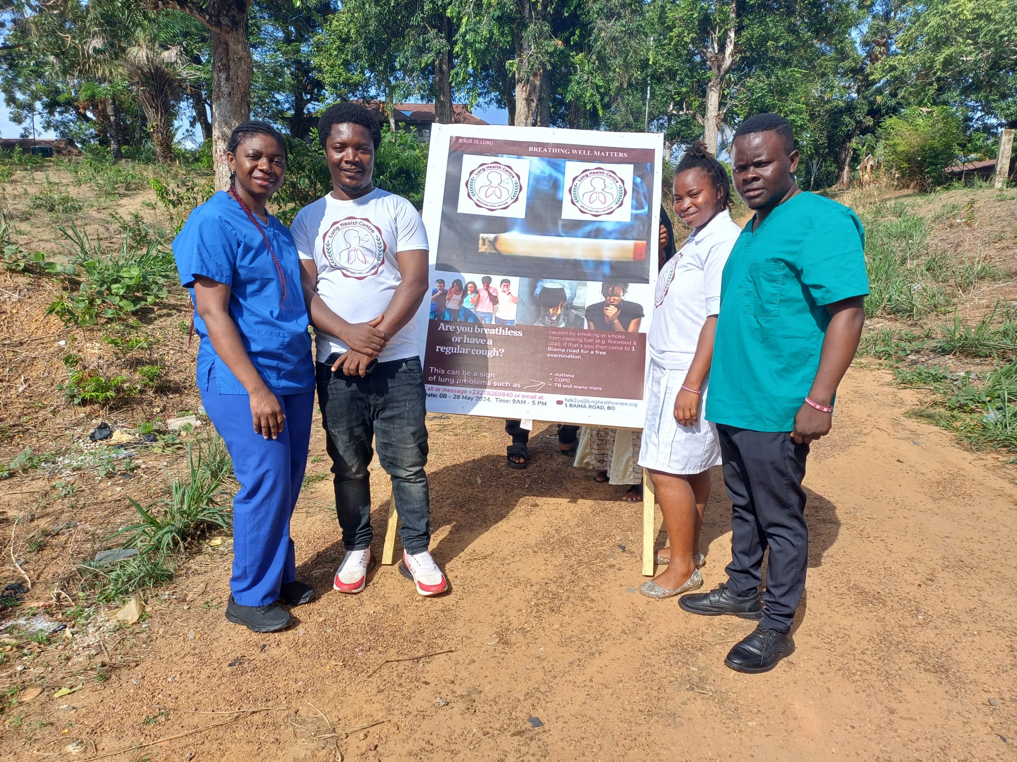 Pilot team in Bo, stood in front of a poster informing people that breathlessness and a persistent cough are symptoms of lung disease.