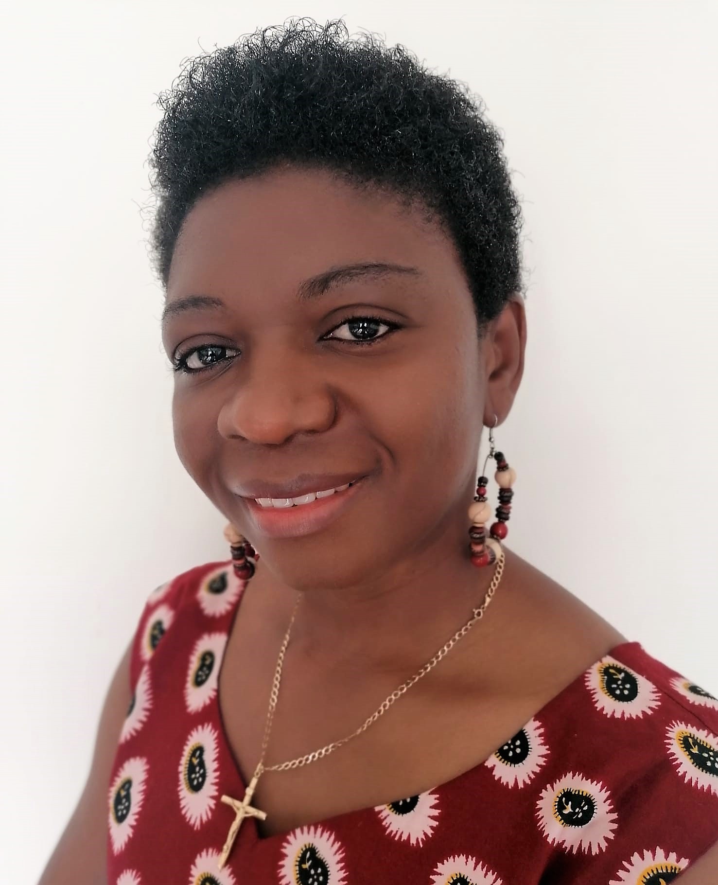 Adama, a black woman with short afro hair is wearing a crucifix necklace, beaded hoop earings and a red shirt with a flower pattern as she smiles at the camera.