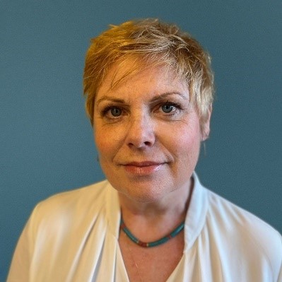 Georgina, a white women with short, cropped blonde hair is wearing a white blouse and smiling at the camera.